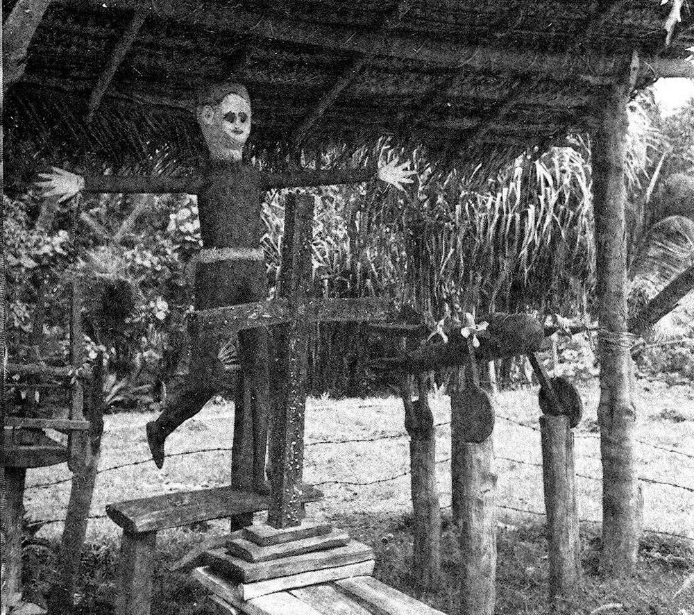 "Symbols of the cargo cult." In the center, a representation of John Frum with "scarlet coat and a white European face" stands behind a brilliantly painted cross. A wooden airplane is on the right, while on the left (outside the photo) a cage contains a winged rat. From Journeys to the Past, which describes Attenborough's visit to the island of Tanna.