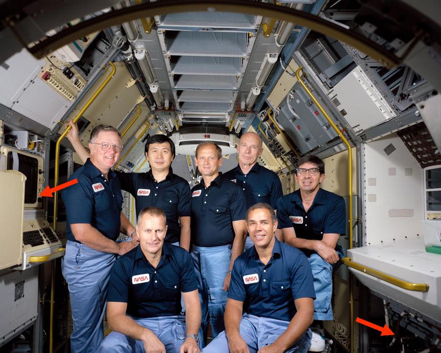 This photo shows astronauts inside Spacelab (but not in space). The Spacelab computers were mounted under the Work Bench (right arrow). The Data Display System (left arrow) provided the interface to the computers. Photo is STS-51B Crew Portrait, 1984.
