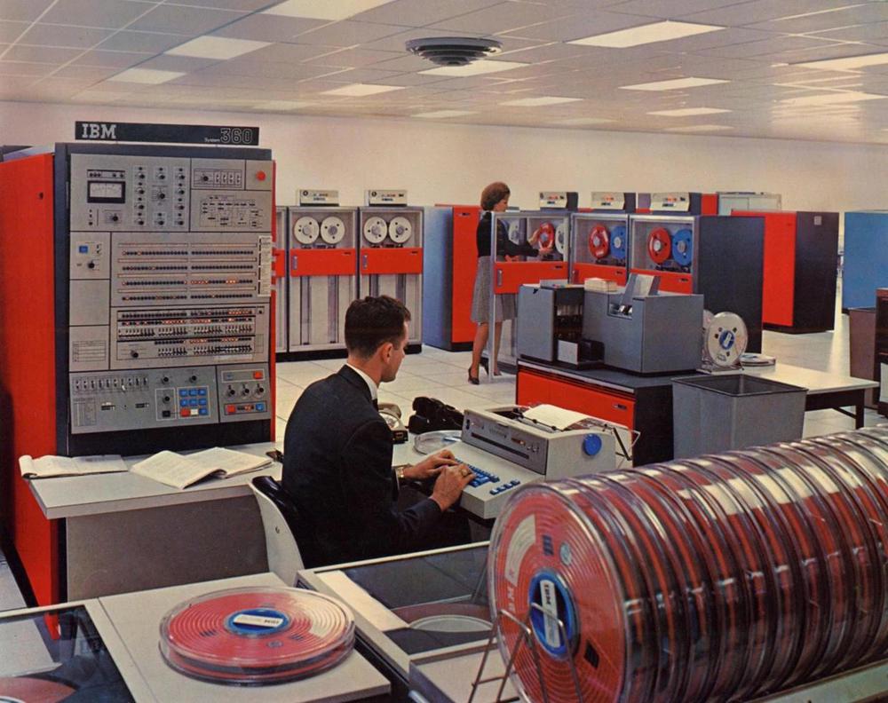 The System/360 Model 50 in a datacenter. The console and processor are at the left. An IBM 1442 card reader/punch is behind the IBM 1052 printer-keyboard that the operator is using. At the back, another operator is loading a tape onto an IBM 2401 tape drive. Photo from IBM.