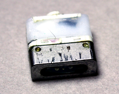 A Magsafe connector with the plastic case removed. In front is the metal holder of the pins. Behind it is the circuit board encased in plastic. The power cable exits from the back.
